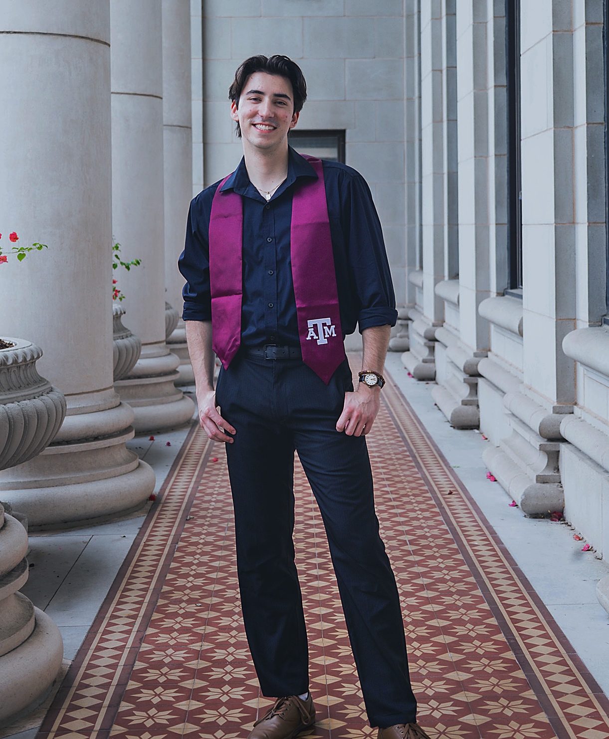 Schultz '25 wearing his maroon Texas A&M graduation stole, standing outside on a patio with columns and a mosaic tile floor. 